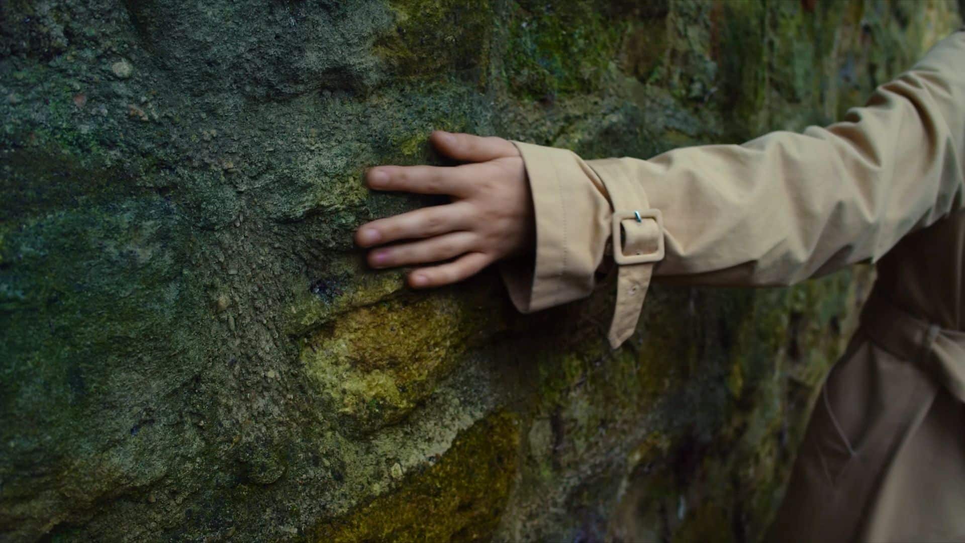 a still from a video. Image shows someone running their hand along a rock with green colouring