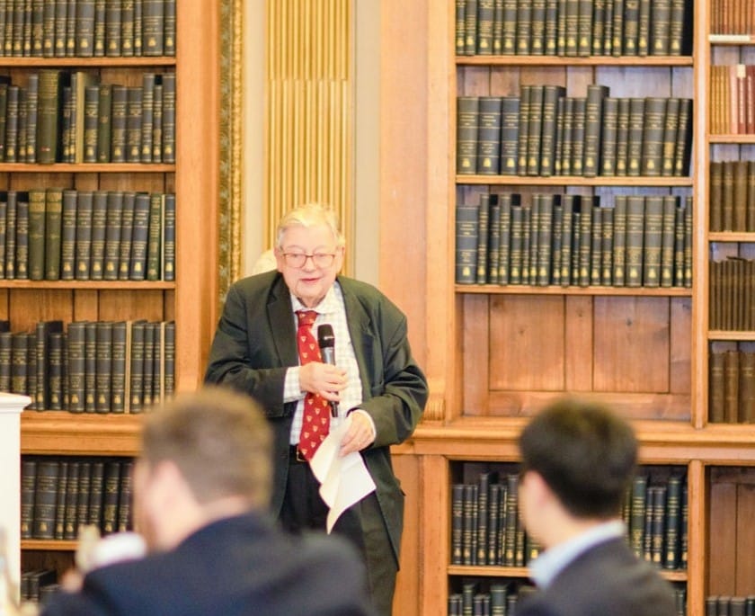 A man with white hair wearing a tie and blazer speaking into a microphone in front of an audience